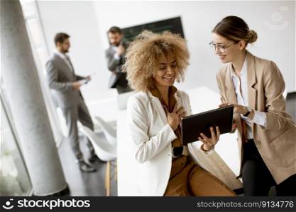 Two young businesswomen discussing with digital tablet in the office with young people works behind them