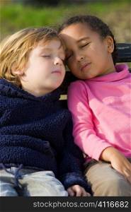 Two young children, one a blond boy the other a mixed race little girl, sleeping on a garden bench.