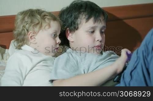 Two young children playing with a tablet computer as they relax together on a sofa with the older brother surfing the internet