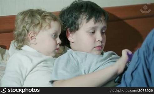 Two young children playing with a tablet computer as they relax together on a sofa with the older brother surfing the internet