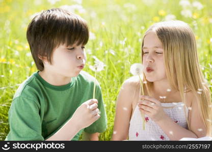 Two young children sitting outdoors blowing dandelion heads smiling