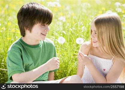 Two young children sitting outdoors holding dandelion heads smiling