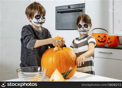 Two young children with skull face paint carving a pumpkin in their kitchen.