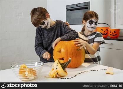 Two young children with skull facepaint cutting a pumpkin in their kitchen.