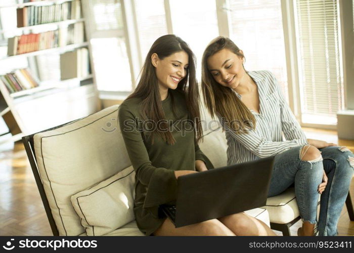 Two young female friends sitting in a room on the sofa and using laptop