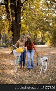 Two young female friends walking in the yellow autumn park with dog and bicycle
