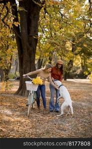 Two young female friends walking in the yellow autumn park with dog and bicycle