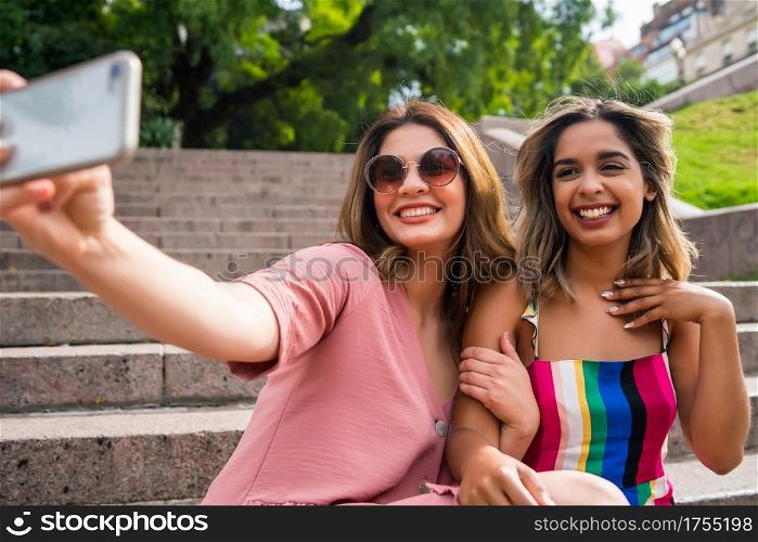 Two young friends having fun together and taking a selfie with their mobile phone while sitting outdoors. Urban concept.