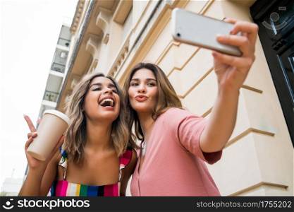 Two young friends having fun together and taking a selfie with their mobile phone while standing outdoors. Urban concept.