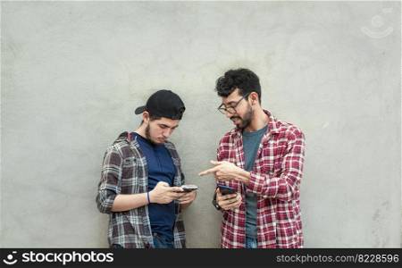 Two young friends leaning against a wall checking their cell phones, Two friends leaning against a wall looking at the content on their phones. Friend showing cell phone to his friend outdoors