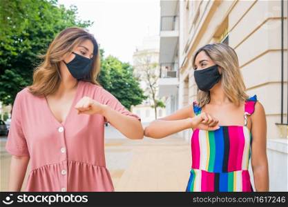 Two young friends wearing face mask and bumping elbows to say hello while standing outdoors. New normal lifestyle concept.