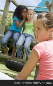 Two young girl friends at a playground whispering about other girl in foreground