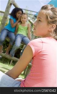 Two young girl friends at a playground whispering about other girl in foreground