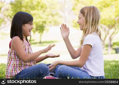Two young girl friends sitting outdoors playing patty cake smiling