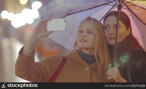 Two young girls friends with multicolored umbrella have a good time while making selfie on the street on rainy day, they talk and laugh