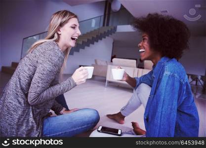 Two young laughing multiethnic women sit on the floor and enjoy while drinking coffee using smartphone in luxury home
