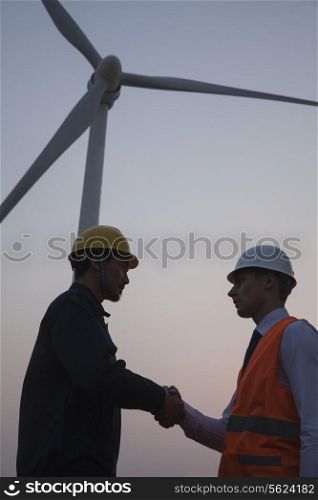 Two young male engineers standing beside a wind turbine at sunset and shaking hands