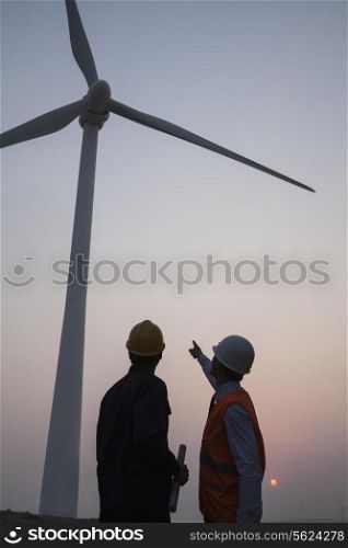 Two young male engineers standing beside a wind turbine at sunset, pointing up