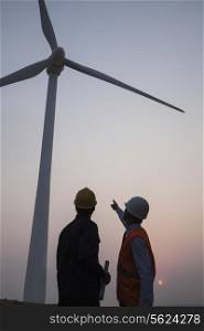 Two young male engineers standing beside a wind turbine at sunset, pointing up