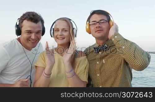 Two young men and woman dancing with music by the sea. Happy people listening favorite rhythms in headphones and getting excited