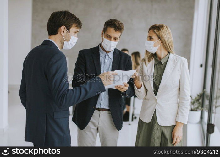 Two young men and woman discussing with paper in hands indoors in the office with young people works behind them