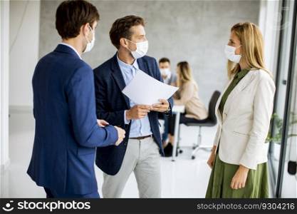 Two young men and woman discussing with paper in hands indoors in the office with young peop≤works behind them