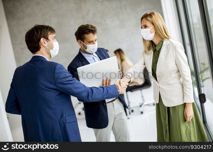 Two young men and woman standing with laptop in hands indoors in the office with young people works behind them