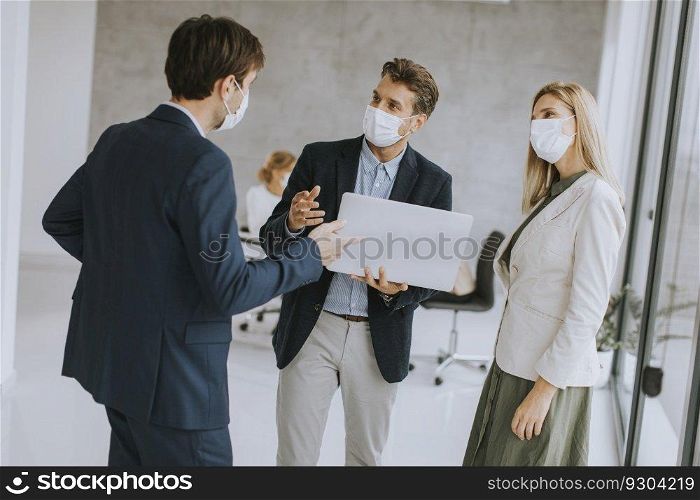 Two young men and woman standing with laptop in hands indoors in the office with young people works behind them