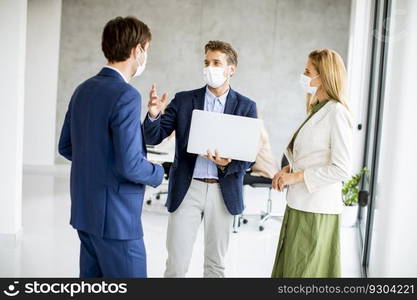 Two young men and woman standing with laptop in hands indoors in the office with young people works behind them