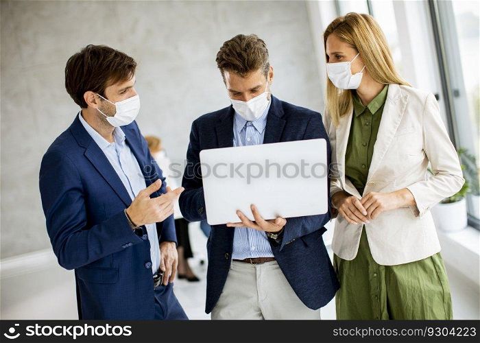 Two young men and woman standing with laptop in hands indoors in the office with young people works behind them