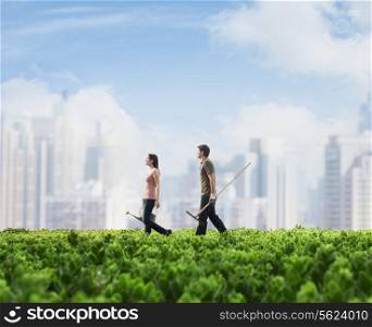 Two young people carrying gardening equipment walking across a green field with plants, cityscape in the background