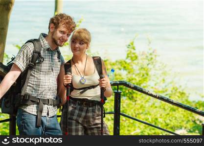Two young people tourists hiking by sea ocean water. Backpackers couple on summer vacation trip journey. Woman with trekking poles sticks.. Two people tourists hiking by sea ocean.