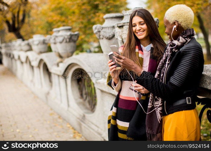 Two young pretty multiracial girlfriends drinking coffee and using mobile phone at autumn day