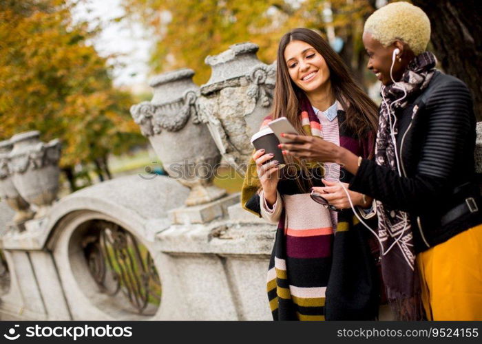 Two young pretty multiracial girlfriends drinking coffee and using mobile phone at autumn day