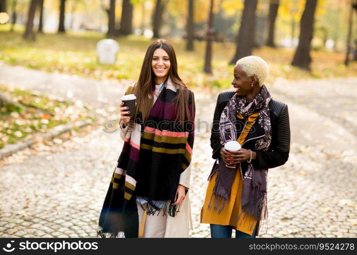 Two young pretty multiracial girlfriends drinking coffee and using mobile phone at autumn day