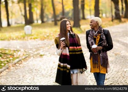 Two young pretty multiracial girlfriends drinking coffee and using mobile phone at autumn day