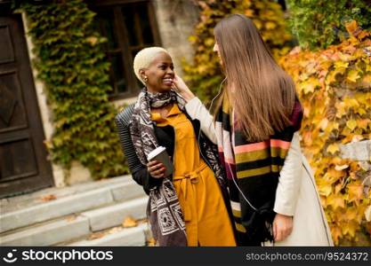 Two young pretty multiracial girlfriends drinking coffee and using mobile phone at autumn day