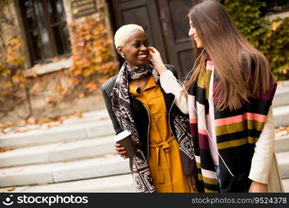 Two young pretty multiracial girlfriends drinking coffee and using mobile phone at autumn day