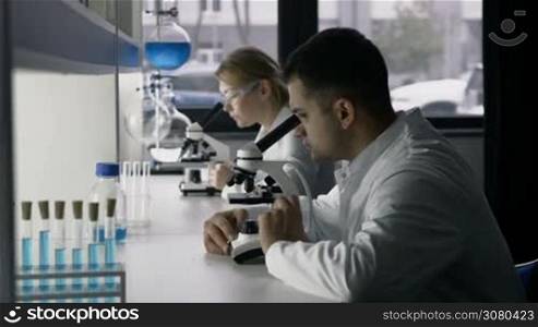 Two young research scientists looking through microscopes studying new substance or virus in modern laboratory. Handsome young mixed race scientist conducting experiment in lab while his female coworker analyzing biological sample under microscope.
