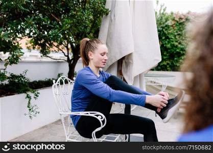 Two young sportswomen tying her shoelaces before the training on a terrace wearing sweatshirt.