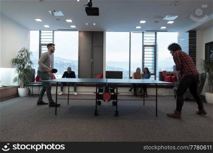 Two young start-up businesses man playing table tennis at modern creative office space people group have meeting and brainstorming in background. High-quality photo. Two young start up business man playing table tennis at modern creative office space people group have meeting and brainstorming in background