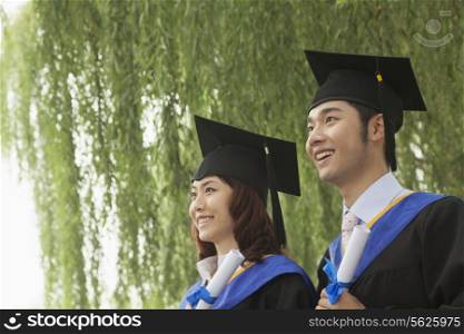 Two Young University Graduates Holding Diplomas And Looking Away
