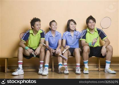 Two young women and two young men sitting side by side on a bench and holding badminton rackets