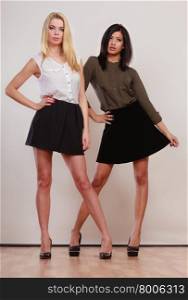 Two young women caucasian and african in trendy short black skirts posing in full length studio portrait on gray