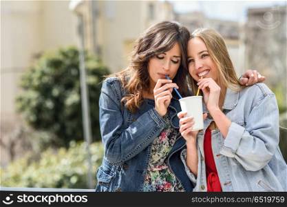 Two young women drinking the same drink with two straws. Two young women drinking the same take away glass together with two straws outdoors.