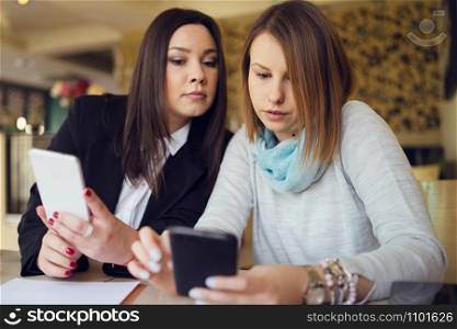 Two young women female friends using smart phone smartphone at the restaurant cafe sitting by the table reading messages or browsing internet watching video or working shopping online