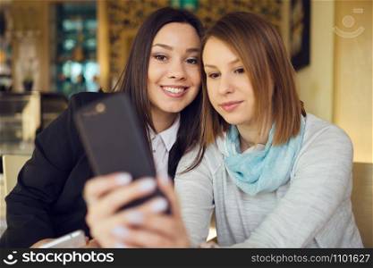Two young women female friends using smart phone smartphone at the restaurant cafe sitting by the table video chat call or taking selfie photos photographs