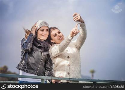Two young women friends using touchscreen for smartphone selfie at coast