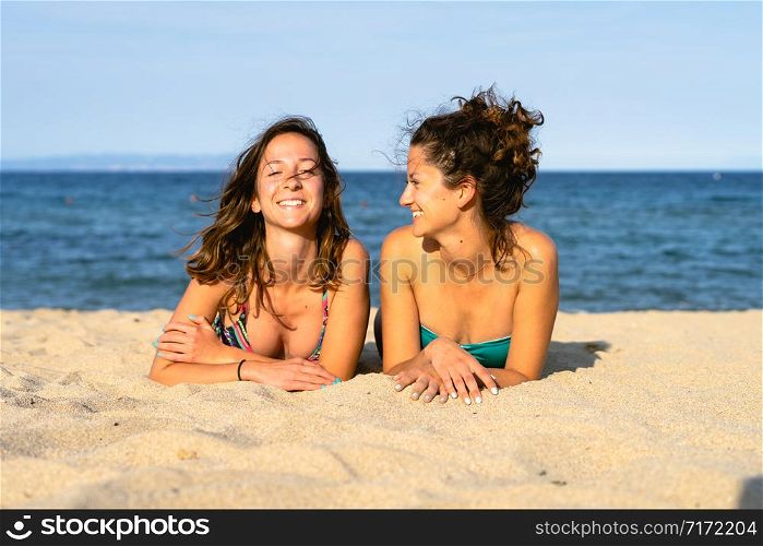 Two young women girl friends or sisters laying on the sand beach by the sea or ocean having fun in summer day sunny sun tanning wearing bikini swimsuit happy tourist