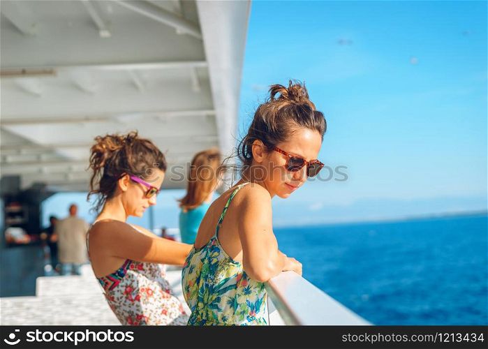 Two young women girl friends sisters standing by the fence on deck of the ferry boat or ship sailing to the island tourist destination on summer vacation looking to the sea in sunny day